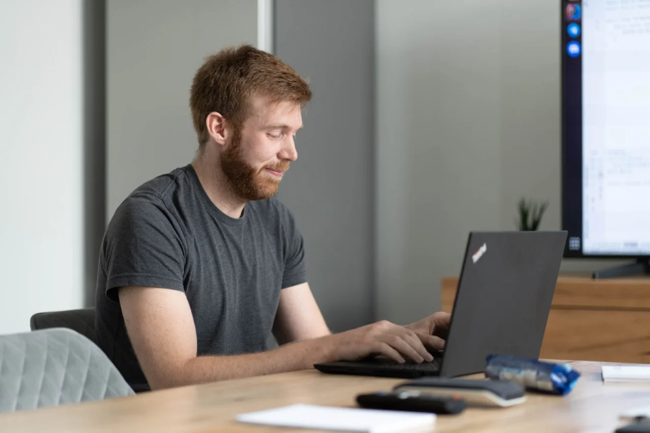 Mann mit Bart sitzt an einem Tisch und tippt auf einem aufgeklappten Laptop.