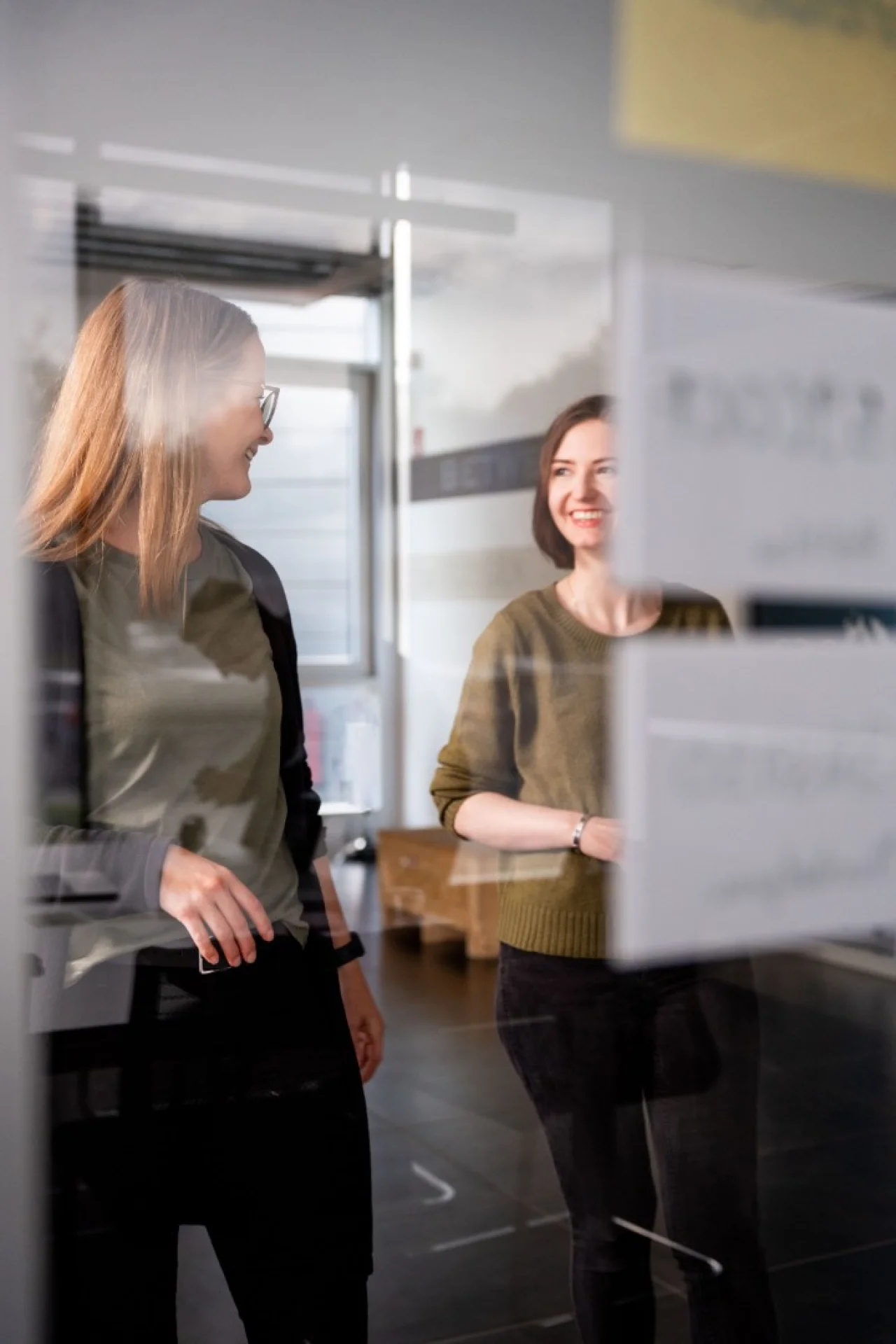 Zwei Frauen unterhalten sich lächelnd in einem modernen Büro, gesehen durch eine unscharfe Glasscheibe mit Post-its.