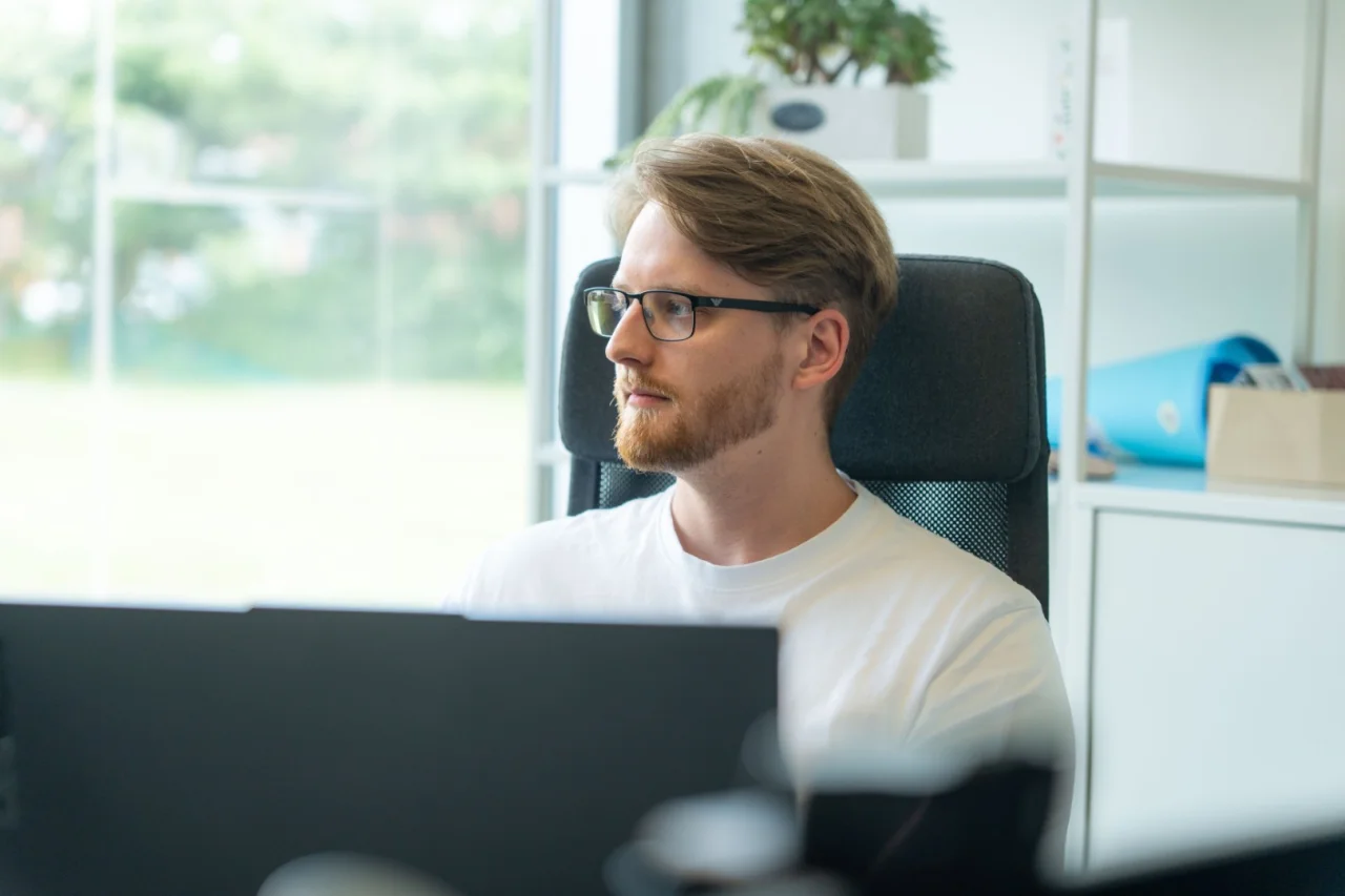 Ein junger Mann mit Brille und Bart sitzt im Büro am Schreibtisch und blickt auf seinen Monitor.