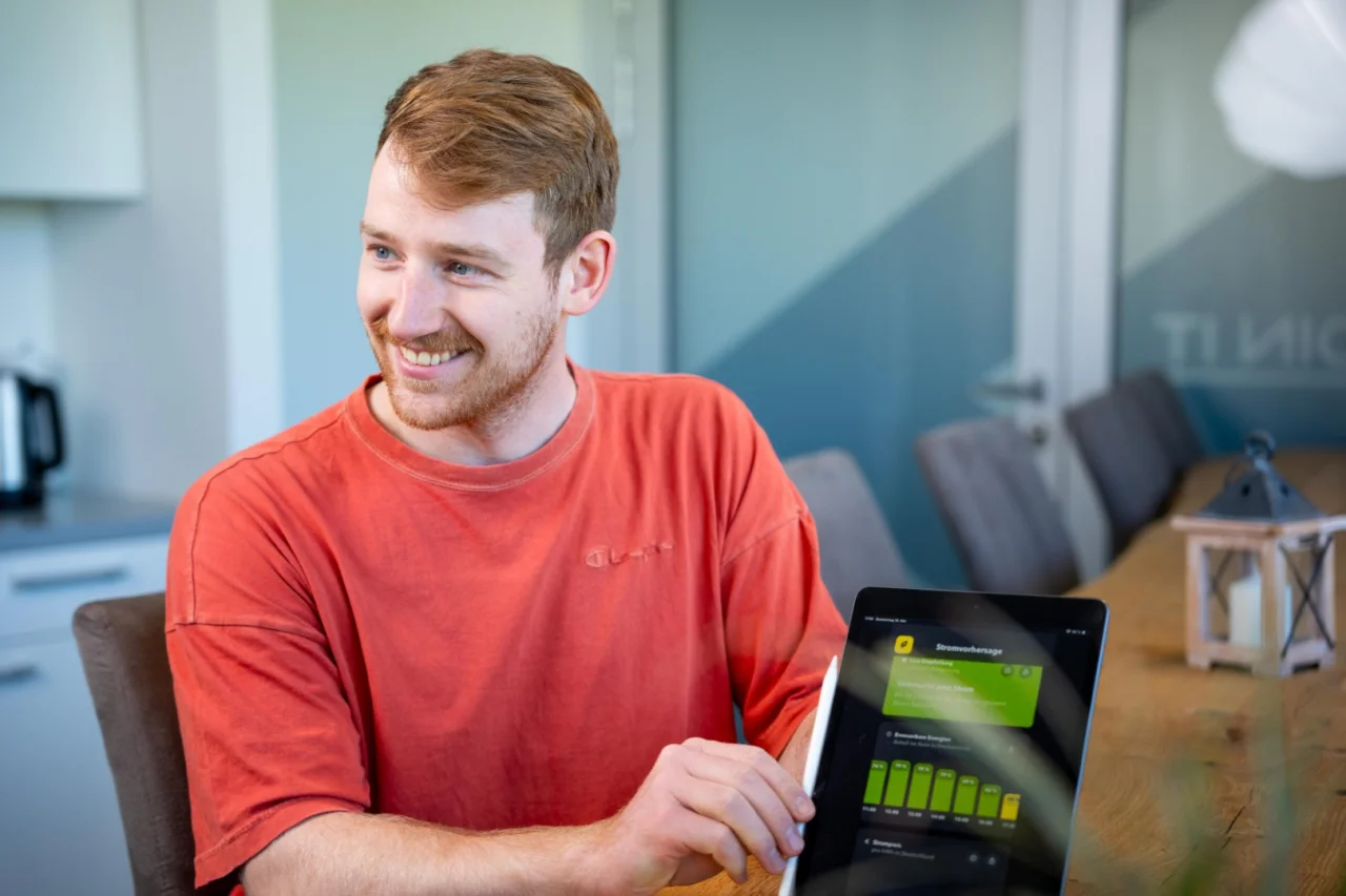 Junger Mann mit rotem T-Shirt sitzt lächelnd an einem Tisch und zeigt auf ein Tablet mit einem Balkendiagramm.