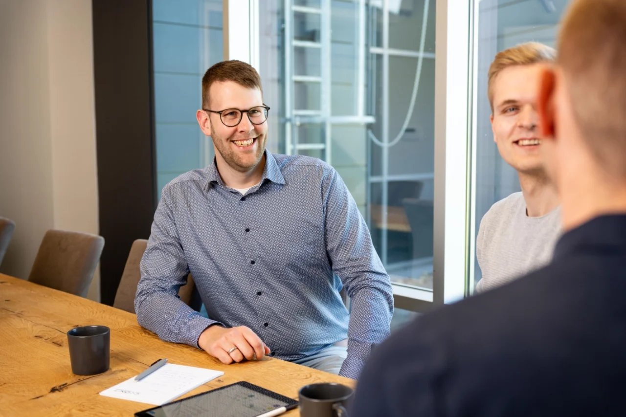 Ein lächelnder Mann mit Brille und blauem Hemd sitzt bei einem Meeting an einem großen Holztisch im Büro.