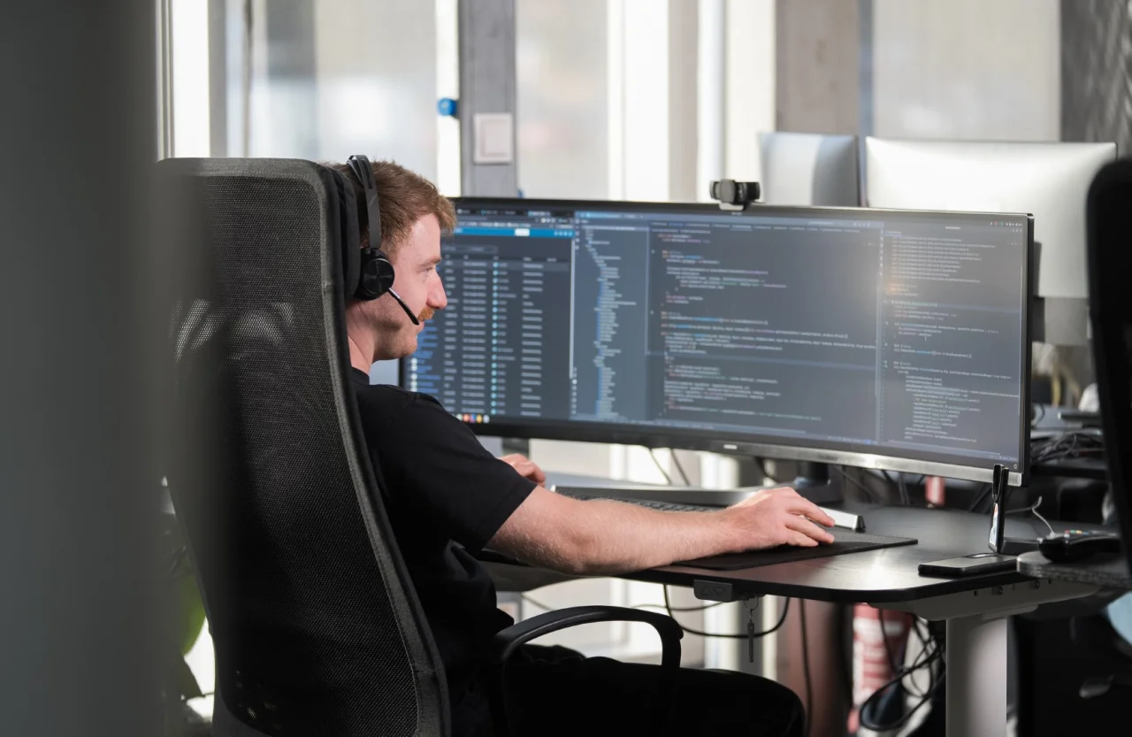 Ein Mann mit Headset sitzt am Schreibtisch und arbeitet vor einem breiten Monitor mit Programmcode.