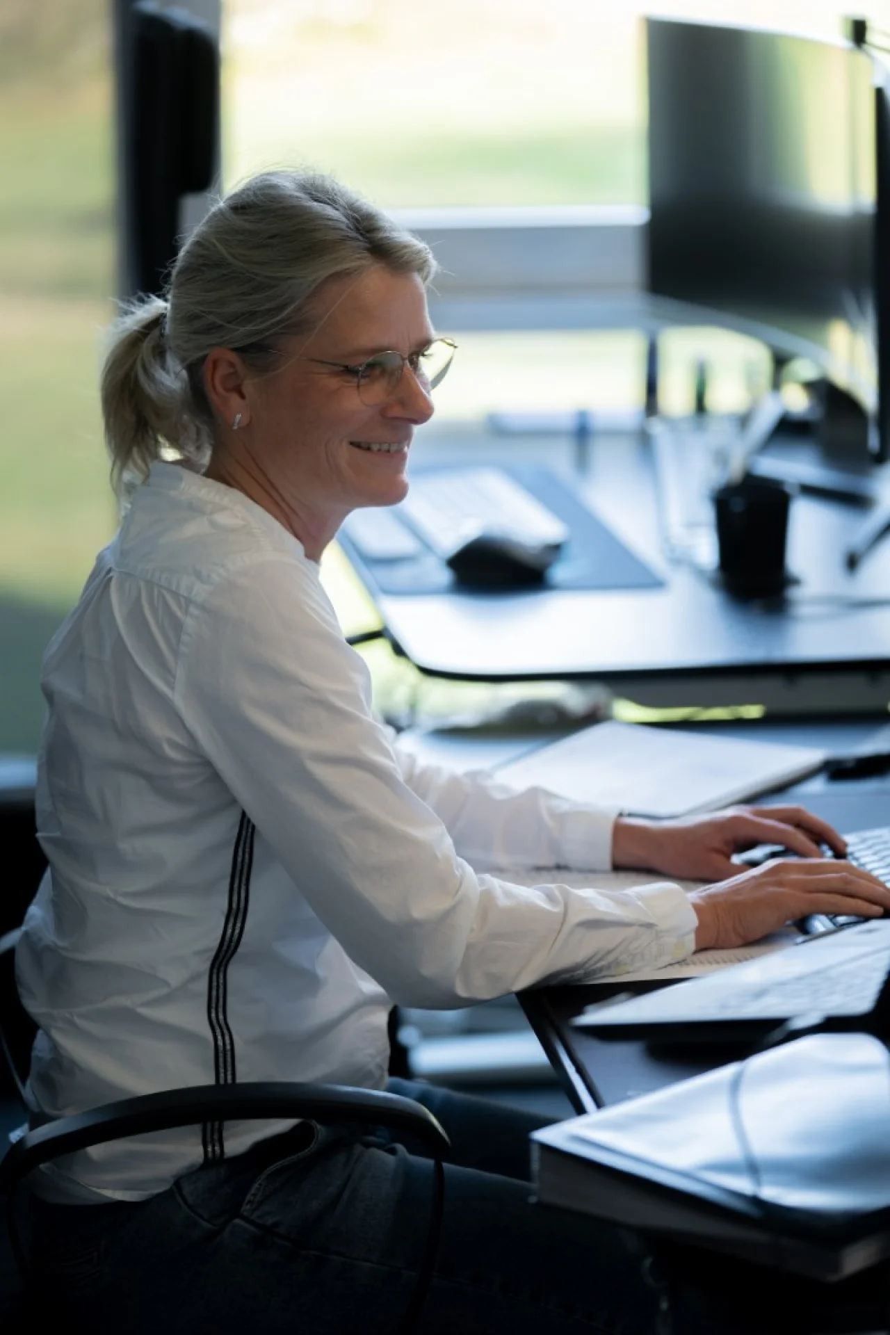 Eine lächelnde Frau mit Brille und weißer Bluse sitzt im Profil an einem Schreibtisch und tippt auf einer Tastatur.