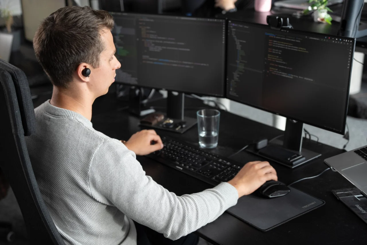 Ein Mann mit In-Ear-Kopfhörer sitzt von hinten an einem Schreibtisch mit zwei Monitoren, auf denen Code angezeigt wird.