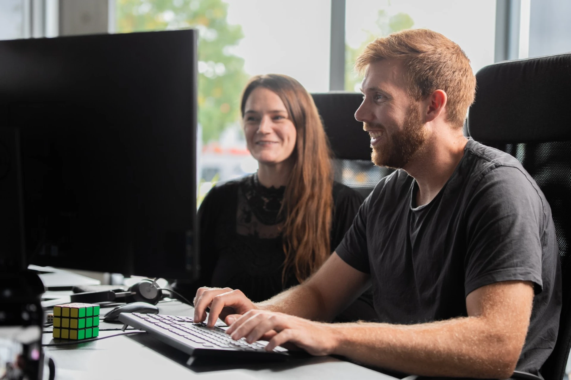 Ein Mann mit Bart sitzt lächelnd vor einem Computerbildschirm, daneben eine Frau mit langen Haaren, auf dem Tisch liegt ein Zauberwürfel.