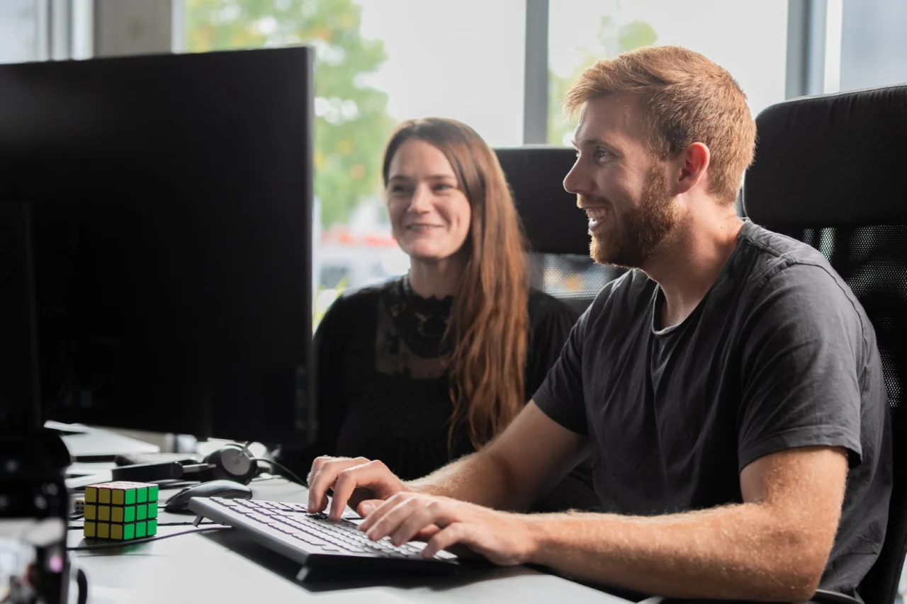 Ein Mann mit Bart sitzt lächelnd vor einem Computerbildschirm, daneben eine Frau mit langen Haaren, auf dem Tisch liegt ein Zauberwürfel.