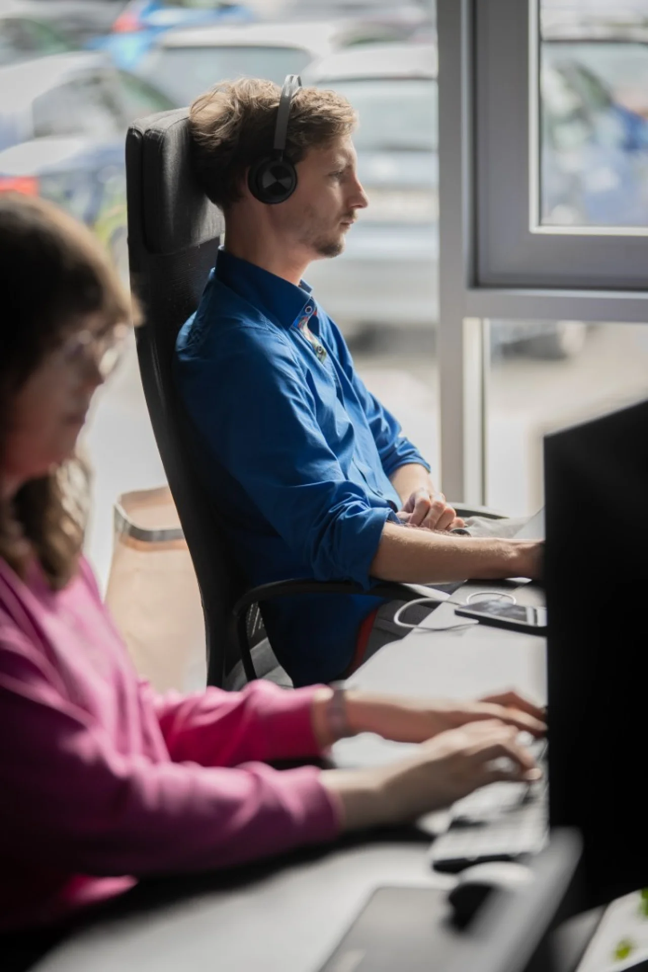 Ein Mann mit Kopfhörern im blauen Hemd sitzt im Profil am Schreibtisch neben einer Kollegin im Büro.
