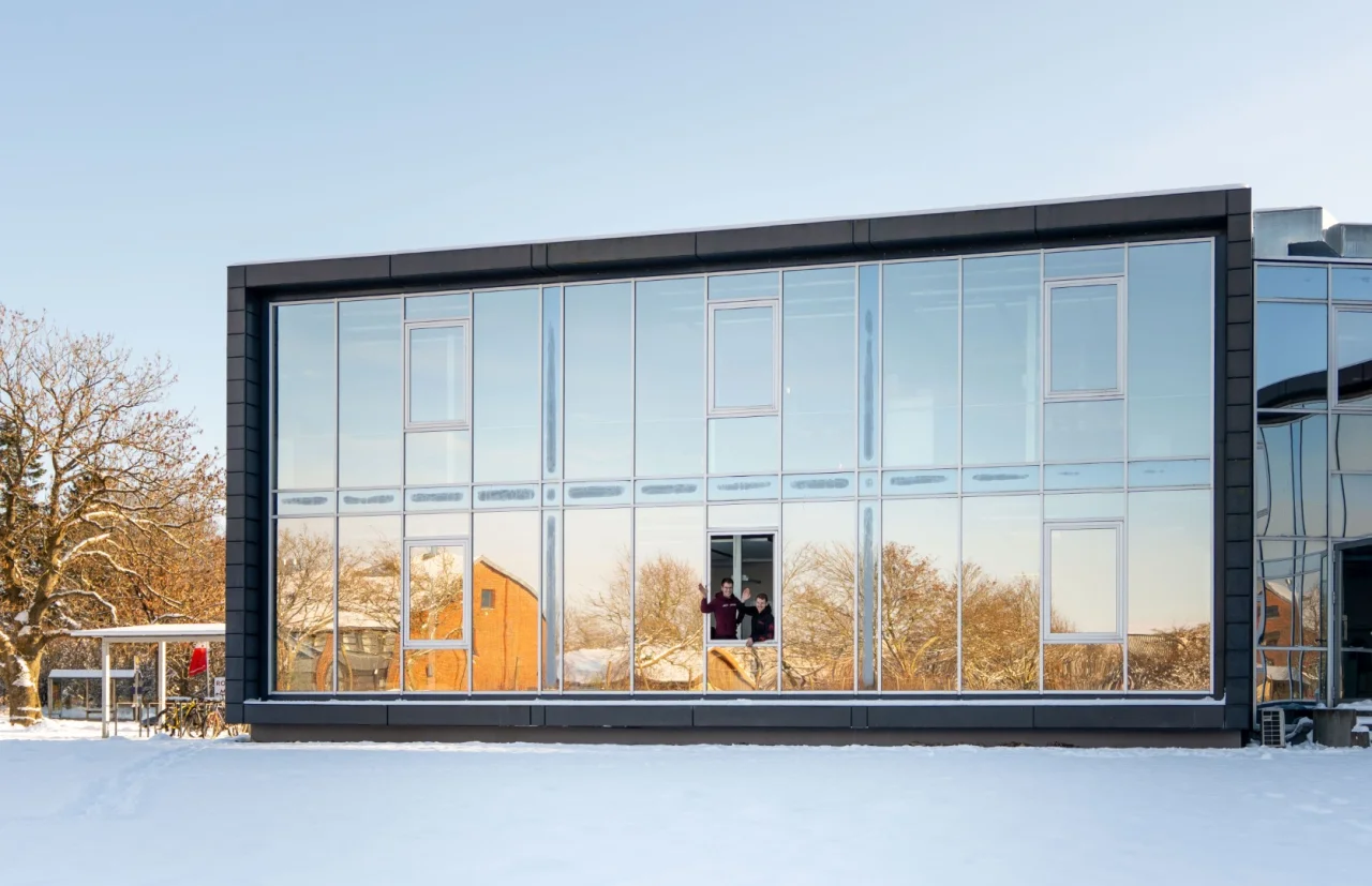 Ein modernes Bürogebäude mit großer Glasfront im Winter, zwei Personen winken aus einem offenen Fenster.