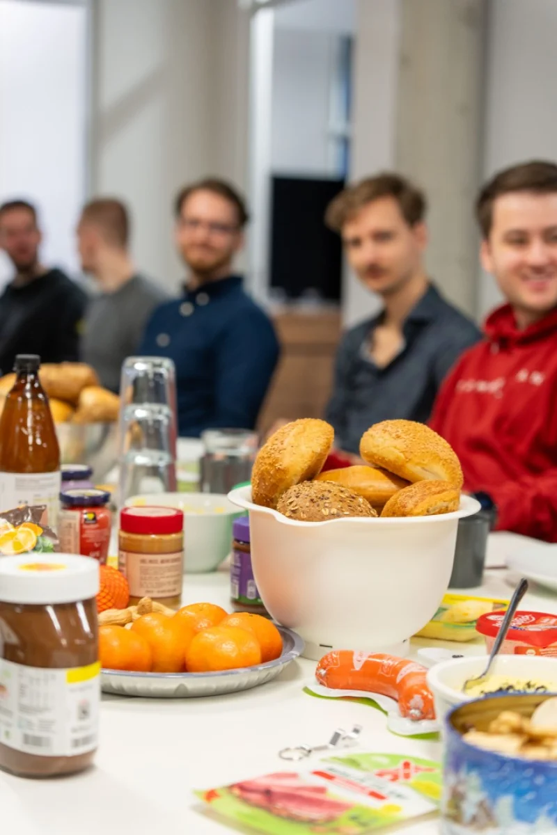 Reich gedeckter Frühstückstisch mit Brötchen und Aufstrichen, im Hintergrund sitzen mehreren Personen.