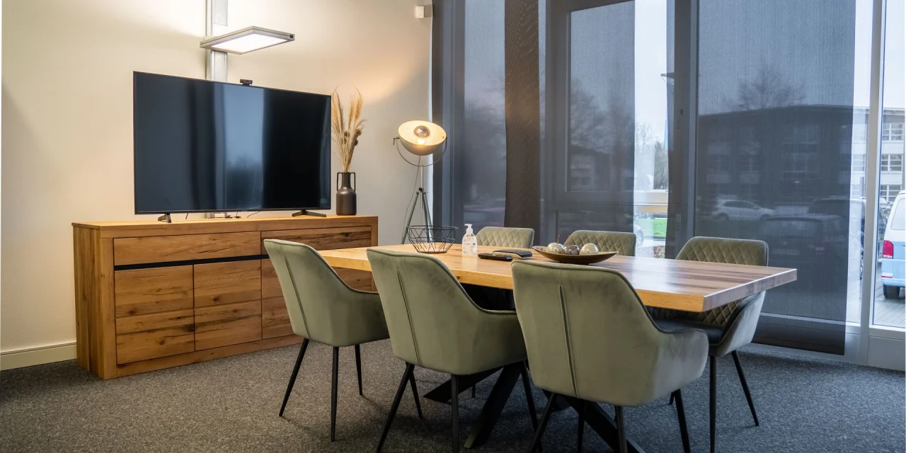 Moderner Konferenzraum mit einem großen Holztisch, grünen Stühlen und einem Fernseher an der Wand.