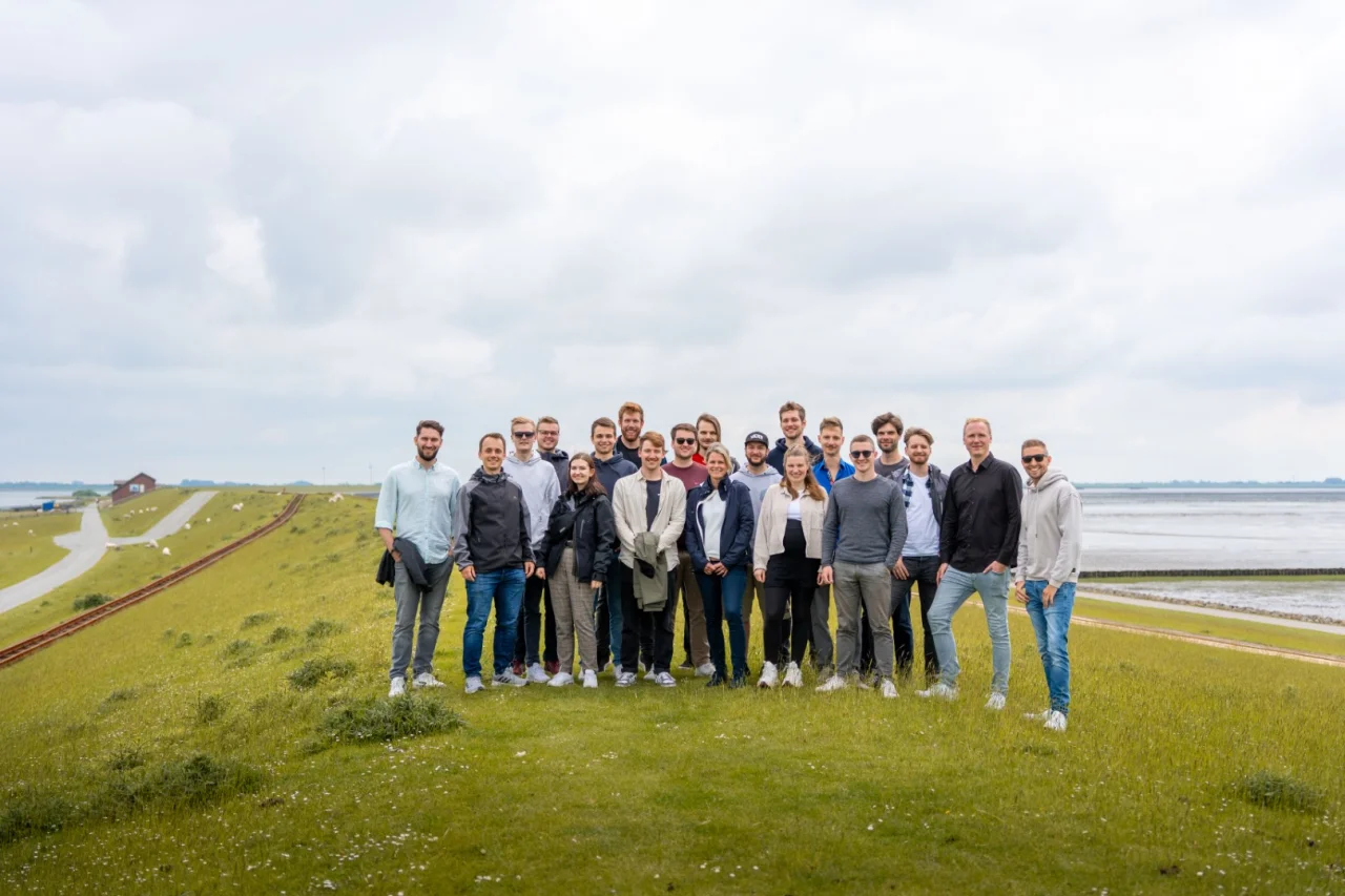 Eine große Gruppe von Menschen steht gemeinsam auf einem grünen Deich unter bewölktem Himmel.