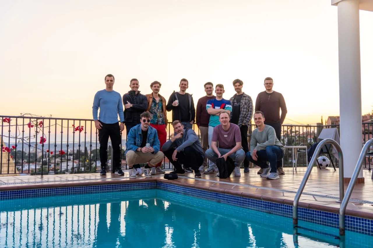 Eine Gruppe von Männern posiert am Rande eines Pools vor einer Balustrade bei Sonnenuntergang.