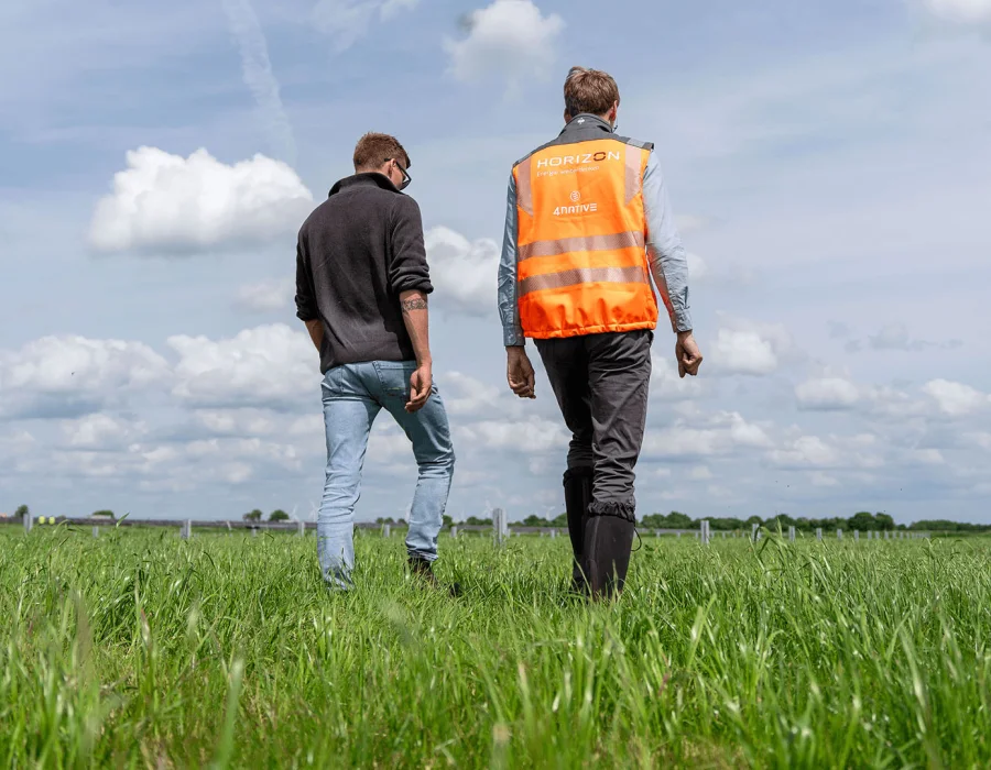 Zwei Männer gehen von hinten gesehen über eine grüne Wiese unter bewölktem Himmel, einer trägt eine Warnweste.