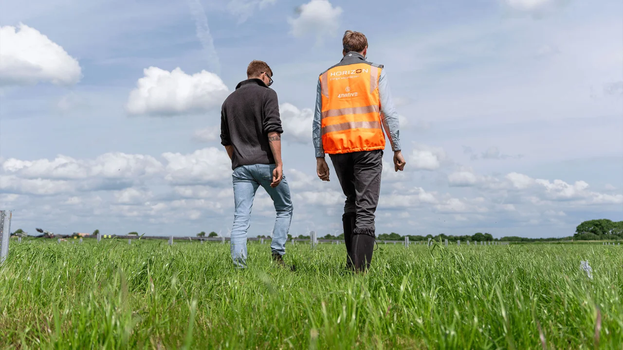 Zwei Männer gehen von hinten gesehen über eine grüne Wiese unter bewölktem Himmel, einer trägt eine Warnweste.