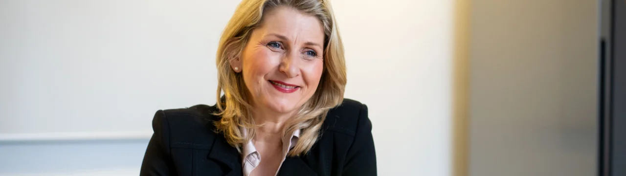 Frau mit blonden Haaren in schwarzem Blazer lächelnd vor hellem Hintergrund sitzend.