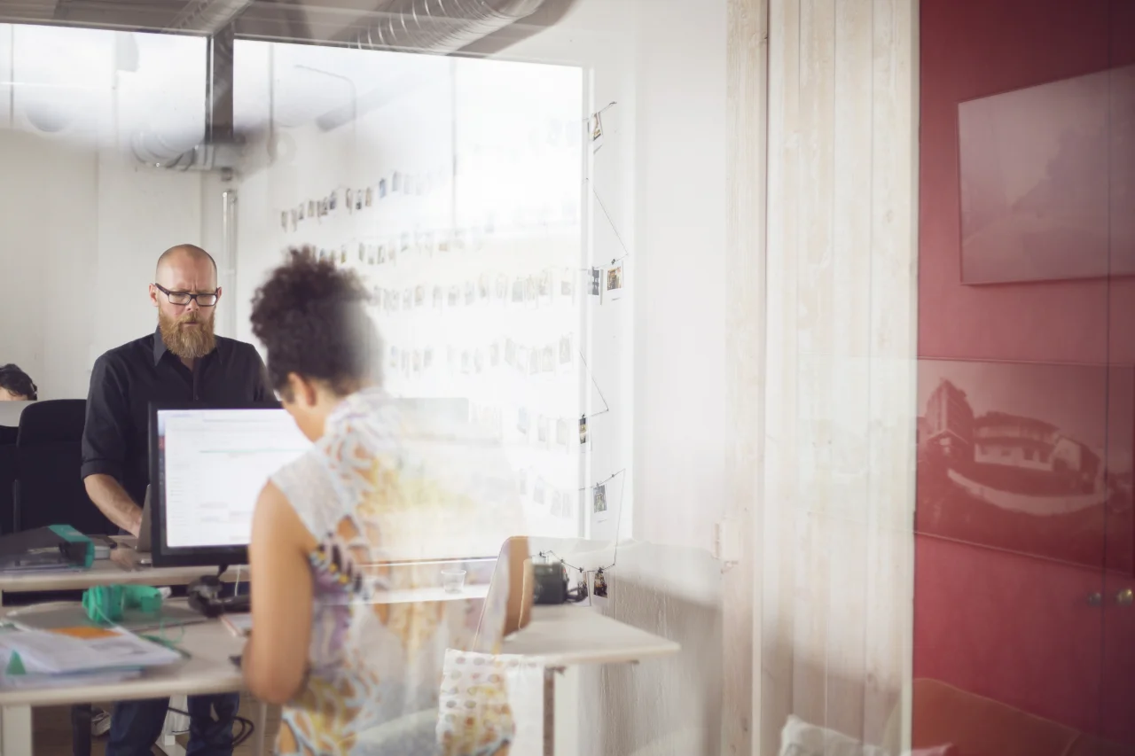 Zwei Personen arbeiten konzentriert an ihren Bildschirmarbeitsplätzen in einem hellen Büro.