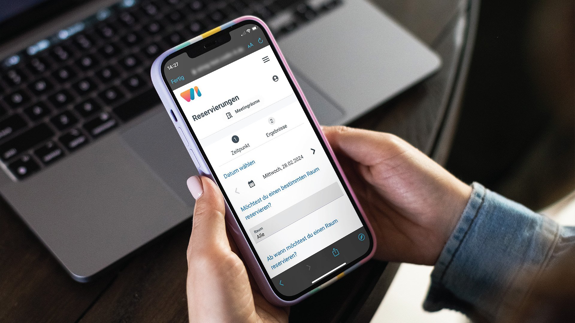 Hände halten ein Smartphone mit einer Reservierungs-App für Meetingräume vor einem geöffneten Laptop.