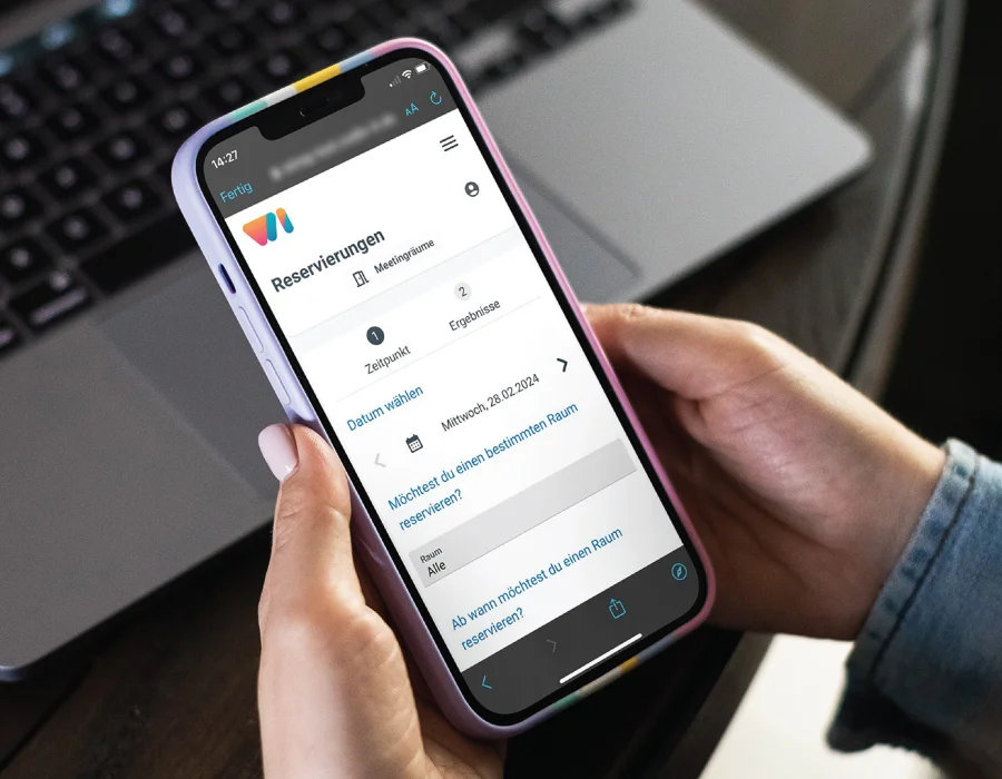 Hände halten ein Smartphone mit einer Reservierungs-App für Meetingräume vor einem geöffneten Laptop.