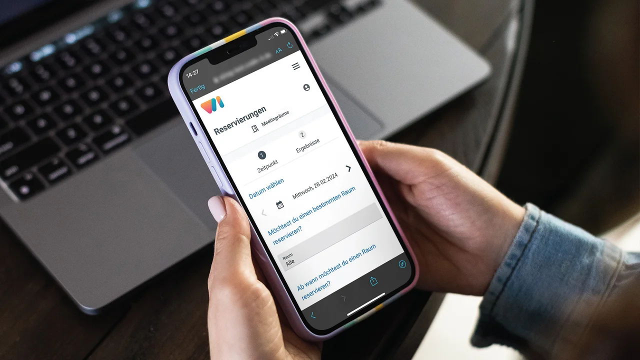 Hände halten ein Smartphone mit einer Reservierungs-App für Meetingräume vor einem geöffneten Laptop.