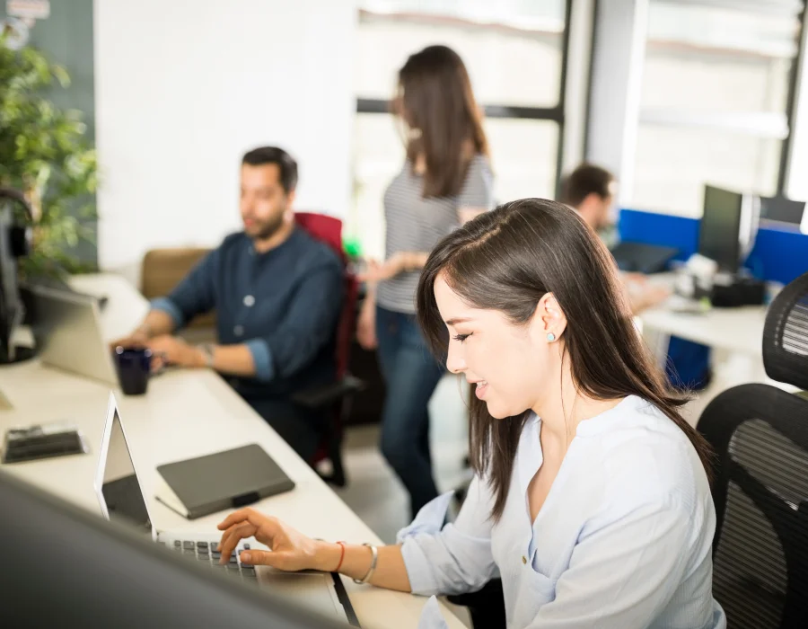 Eine lächelnde Frau tippt auf ihrem Laptop im Büro, während Kolleginnen im Hintergrund arbeiten.