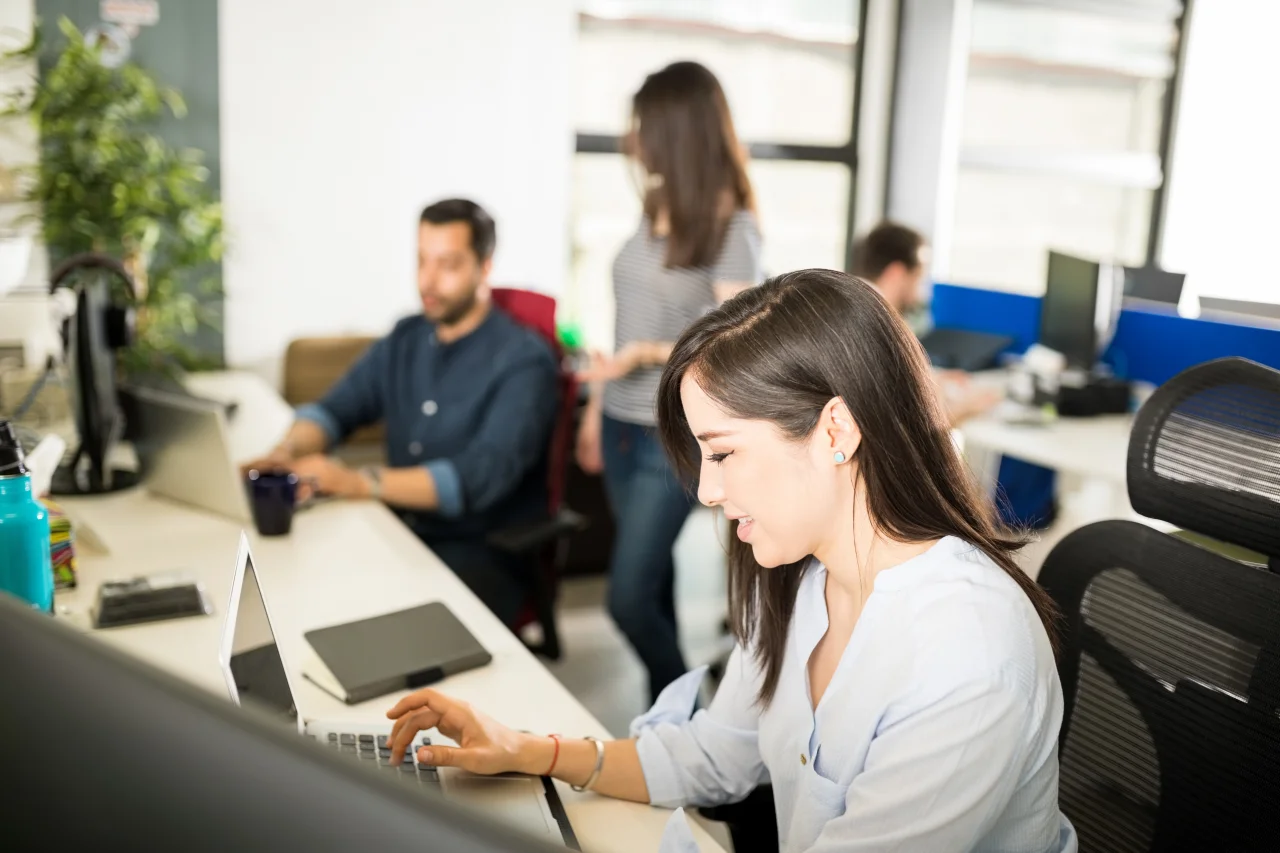 Eine lächelnde Frau tippt auf ihrem Laptop im Büro, während Kolleginnen im Hintergrund arbeiten.